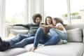 Family reading together on a gray sofa by large windows, with a book in the mother's hands.
