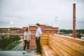 Two people talking on a rooftop with a wooden bench, overlooking a brick building and Lucky Strike smokestack.