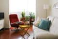 Living room with red swivel chair, beige sofa, colorful pillows, yellow tray table, and potted plants by the window.