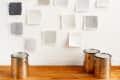 Paint swatches in various shades of gray on a wall above three metal paint cans on a wooden floor.