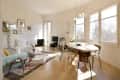 Bright living and dining area with a round table, mixed chairs, sofa, wall art, and large windows overlooking trees.