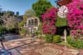 Historic estate courtyard with ornate tile flooring, lush greenery, vibrant pink flowers, and a large vintage clock.