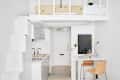 Compact white kitchen with copper fixtures, loft bed above, and a small dining area with woven chairs and a vase on the table.