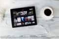 Tablet displaying Netflix interface on a wooden table with a cup of coffee and white earphones.