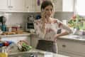 Woman in floral top talking on phone in a kitchen with plants, colorful towels, and baking items on the counter.