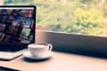Laptop displaying Netflix shows next to a cup of coffee on a windowsill with a blurred green view outside.