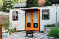 Shipping container home with orange double doors, patio chairs, fire pit, and potted plants in a garden setting.