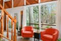 Living room with large windows, two orange armchairs, a small table, and potted plants, overlooking a wooded area.