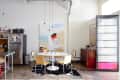 Dining area with round white table, four yellow chairs, abstract wall art, and a stainless steel fridge with photos.