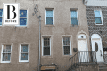 Two-story beige townhouse with four windows, white door, and black railing, next to a brick house.