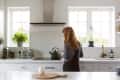 Woman in a modern kitchen with white cabinets, black kettle, toaster, and potted plants on the counter.