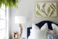 Bedroom with a navy headboard, white bedding, wooden nightstand, lamp, hanging fern, and bird artwork on the wall.