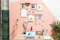 Desk with laptop, clock, and decor against a peach wall with art and plants.