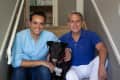 Two men sitting on stairs with a black dog, smiling at the camera.