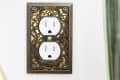 Decorative brass outlet cover with intricate floral and scroll design on a white wall.