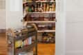 Under-stair pantry with open doors, shelves of jars, cans, and baskets, and a wooden cart with jars and boxes on a wooden floor.