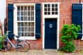 Brick house facade with large window, blue shutters, blue door, bicycle, and green shrub.