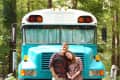 Couple standing in front of a turquoise converted bus in a wooded area.