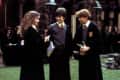 Three students in school uniforms and robes talking in a courtyard, holding books.
