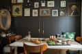 Dining room with dark walls, gallery wall art, white table, wooden chairs, and a spread of food and drinks.