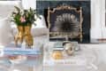 Living room with a white fireplace, abstract painting, gold candlesticks, and a glass coffee table with decor books and plants.