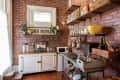 Rustic kitchen with brick walls, open shelves, white cabinets, and various kitchen items including jars, plants, and a yellow bowl.