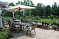 Patio with wooden dining set under a large umbrella, surrounded by lush garden and stone wall with potted plants.