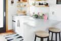 Modern kitchen with white cabinets, open shelves, plants, and wooden stools at a breakfast bar.