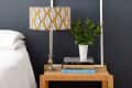Bedside table with a patterned lamp, potted plant, and stacked books against a dark wall.