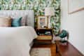 Bedroom with tropical leaf wallpaper, tufted headboard, colorful pillows, wooden nightstand, lamp, and vintage fan.