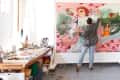 Artist painting a large colorful mural in a studio, surrounded by art supplies on a wooden table.
