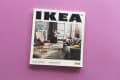 IKEA catalog on a purple surface, featuring a living room with a sofa, armchair, and colorful rug.