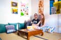 Couple sitting on floor cushions in a room with colorful posters and a wooden coffee table with books.