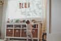 Toddler in striped pants reaching for toys on a shelf under a window with "ELLA" hanging letters.