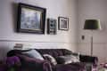 Plum velvet sofa with cushions, framed art on wall, floor lamp, books, and flowers on wooden floor with pink rug.