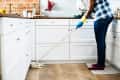 Person mopping a kitchen floor with white cabinets, wearing blue gloves and jeans, near a brick wall and countertop items.