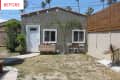 Small backyard with a gray stucco house, white door, string lights, potted plants, and a small patio set.