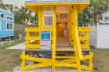 Bright yellow tiny house with front steps, open door, and rip currents safety sign, set in a grassy area with trees.