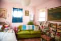 Cozy room with pink walls, green sofa, stacks of books, framed art, and a small dog on the bed.