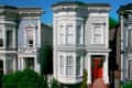 Victorian-style house with red door, ornate white trim, and bay windows, flanked by similar houses.
