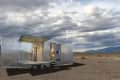 Modern silver trailer with open glass doors in a desert landscape under a cloudy sky.