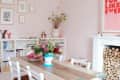 Dining room with a wooden table, white chairs, pink walls, floral centerpiece, and a fireplace filled with logs.
