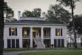 Historic white plantation-style house with large columns, black shutters, and a central staircase, surrounded by trees.
