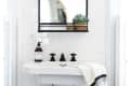 A rectangular mirror in a black frame above a white bathroom sink with black fixtures