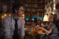Man in glasses and tie sitting on bed with child wearing a shark hat, surrounded by books and warm lighting.