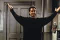 Man in a black sweater with arms raised, standing in front of a closed door.