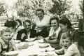 Family gathered around an outdoor table, smiling and serving soup, with trees and a wooden fence in the background.