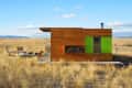 Small wooden cabin with green accent wall, firewood storage, and outdoor seating on a grassy plain under a blue sky.