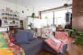 Cozy living room with a gray sofa, colorful pillows, bookshelves, and hanging plants, featuring brick walls and large windows.