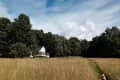 White house with a wraparound porch surrounded by trees, set in a grassy field under a blue sky.